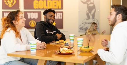 A group of students, one white male, one white female and one black male, seated at a table in a dining hall on campus at Kutztown University laughing and eating