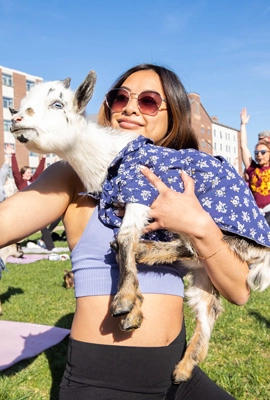 A diverse female holding a goat and taking a selfie on a sunny day outside with others doing yoga in the near background